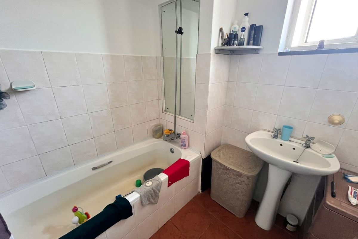 A functional bathroom featuring a bathtub with a shower screen, pedestal basin, and tiled finishes, currently requiring cosmetic updates and maintenance.