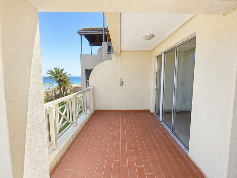 A spacious covered balcony featuring terracotta tiling, a protective railing, and sliding glass door access, offering a pleasant outdoor area with coastal views.