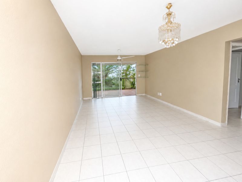 A spacious, empty living room featuring neutral tiled flooring, a ceiling fan, a decorative chandelier, and sliding glass doors leading to an outdoor area.