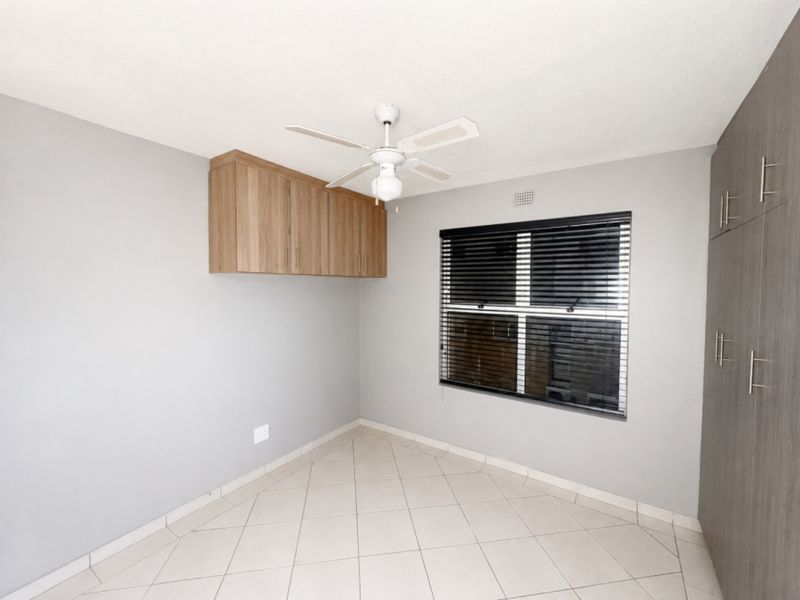 A clean, empty bedroom featuring neutral tiled floors, built-in storage cupboards, wall-mounted cabinetry, and a ceiling fan.