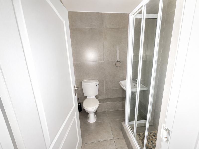 A compact, modern bathroom featuring neutral grey floor-to-ceiling tiling, a white toilet, a wall-mounted basin, and a glass-enclosed shower.