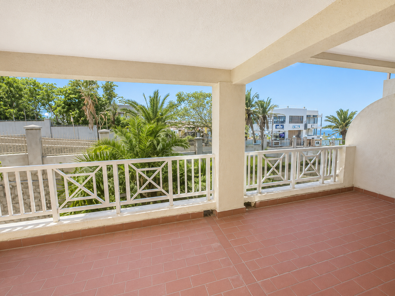 A spacious covered balcony featuring terracotta-style tiled flooring and a white metal balustrade, offering an open outdoor area with views of the surrounding neighborhood.