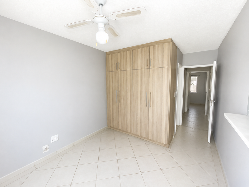 A bright, empty bedroom featuring light-toned built-in wardrobes, neutral tiled flooring, and a ceiling fan, offering a clean and versatile living space.