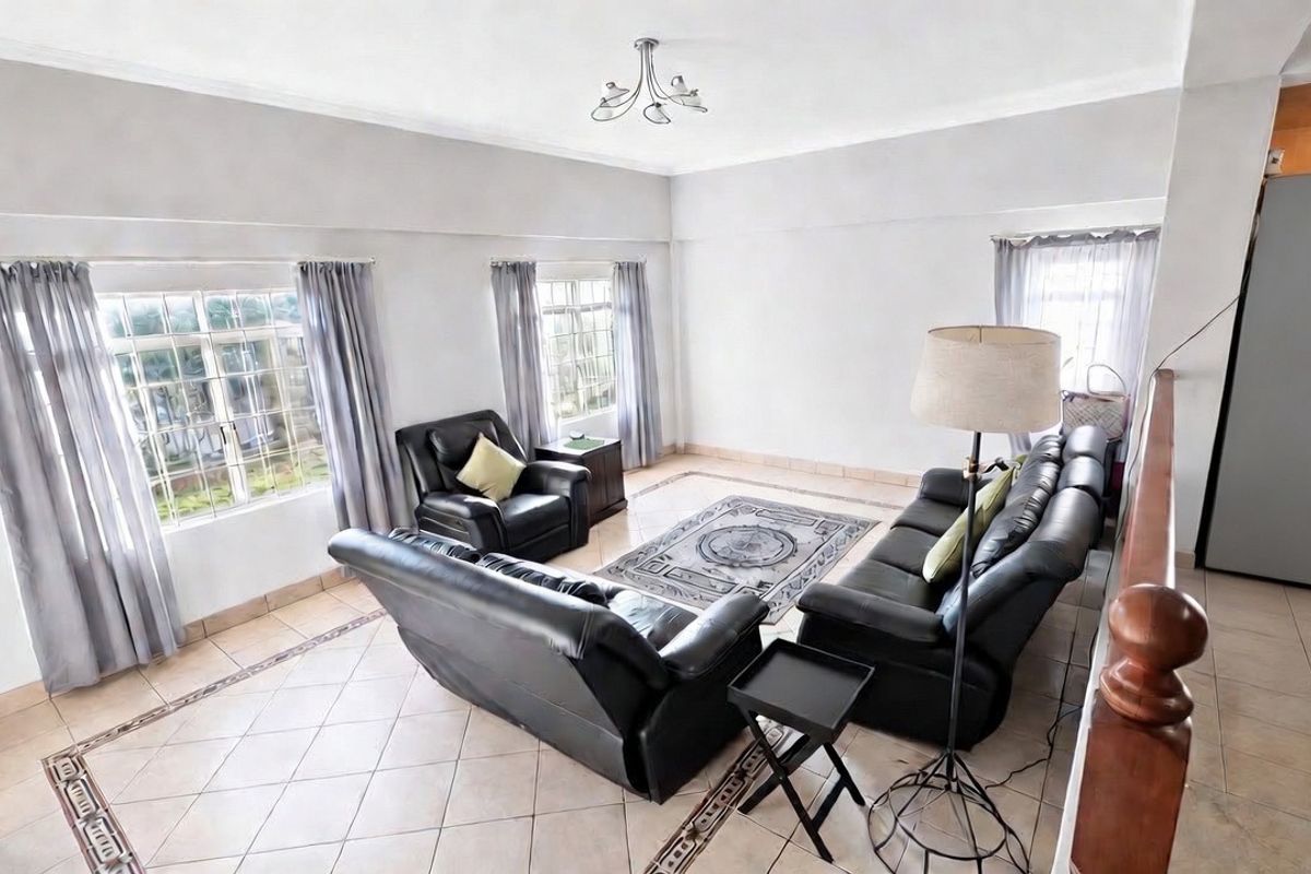 Spacious living room featuring tiled flooring with a decorative border, large windows with security bars, and a wooden staircase railing.