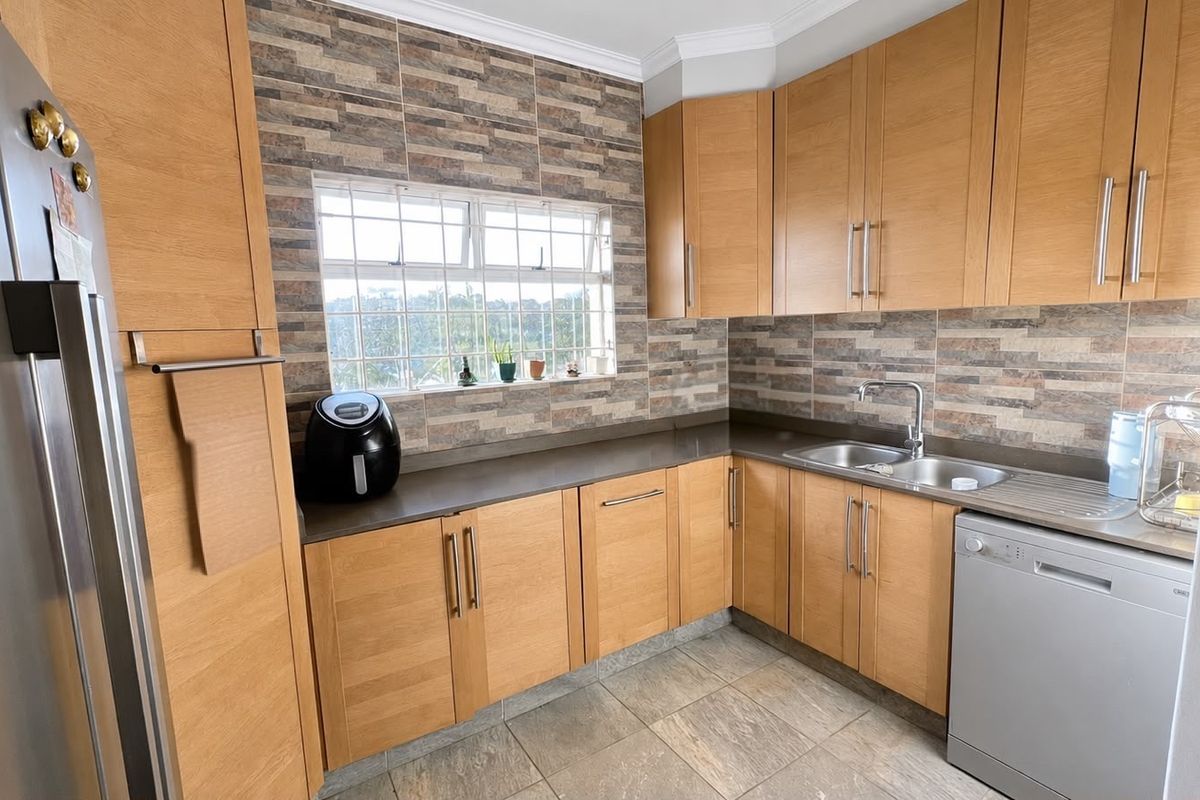 A functional kitchen featuring light wood cabinetry, stone-look tile backsplash, stone countertops, and integrated stainless steel sink with modern fixtures.