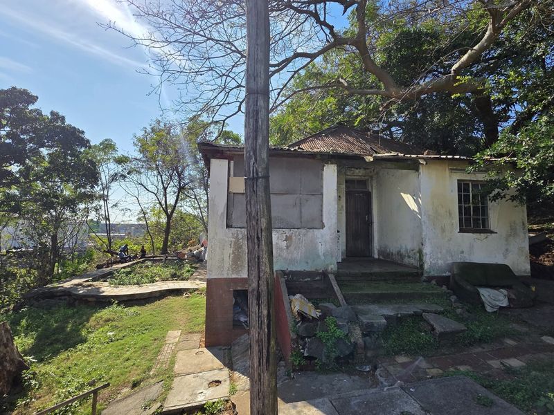 A weathered single-story residential structure featuring a corrugated iron roof and masonry walls, situated on a sloped plot with mature trees.