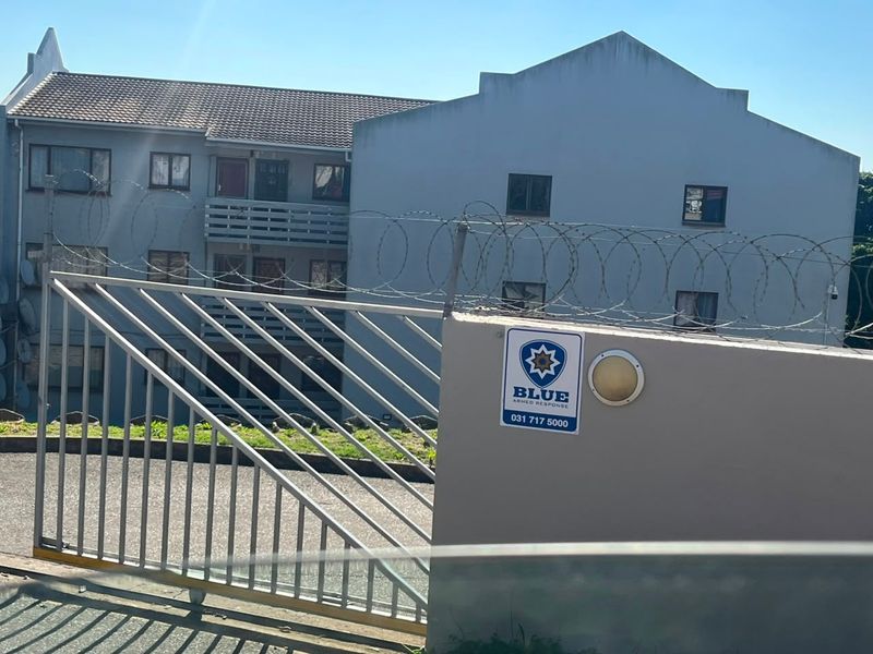 Exterior view of an apartment complex featuring a perimeter wall with razor wire, a security gate, and multi-story residential units with balconies.