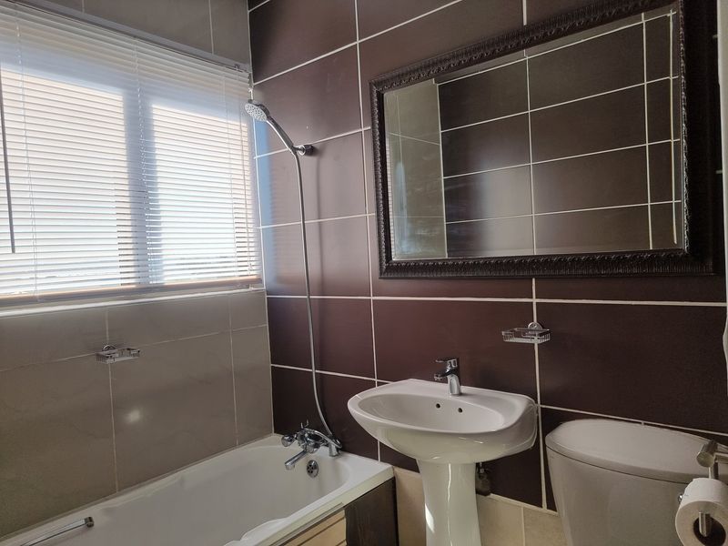 A clean bathroom featuring a pedestal basin, bathtub with shower mixer, and dark wall tiling with a window fitted with venetian blinds.