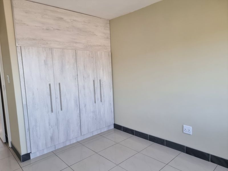 A clean, empty bedroom featuring light wood-grain built-in cupboards, neutral wall paint, and tiled flooring with dark skirting boards.
