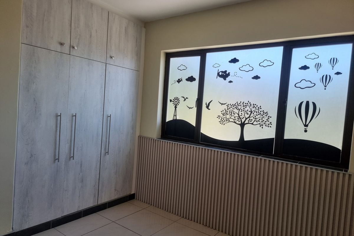 A clean, empty bedroom featuring modern light wood-grain built-in cupboards, tiled flooring, and decorative slatted wall paneling beneath a window with privacy film.