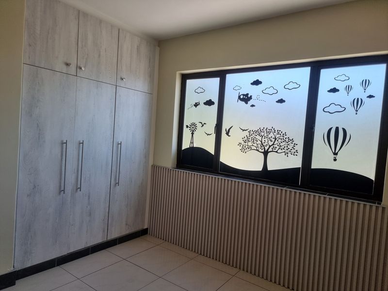 A clean, empty bedroom featuring modern light wood-grain built-in cupboards, tiled flooring, and decorative slatted wall paneling beneath a window with privacy film.
