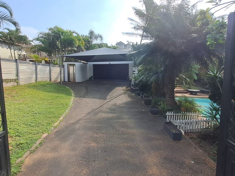 Front entrance view featuring an asphalt driveway, shade sail carport, garage, and a swimming pool area enclosed by a perimeter wall.