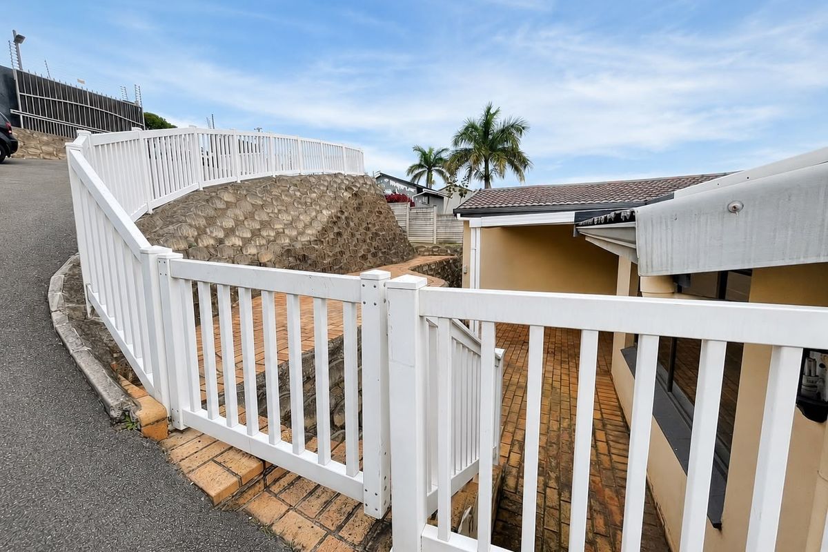 Rear exterior view featuring a paved walkway, retaining wall, and white vinyl fencing leading towards the side of the house.