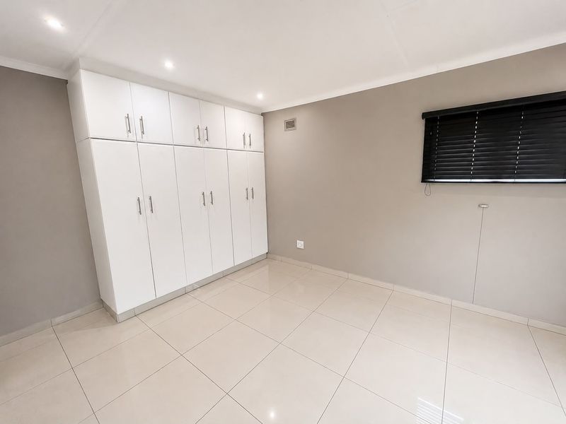A clean, empty bedroom featuring neutral tiled flooring, modern built-in cupboards, recessed ceiling lights, and dark venetian blinds.