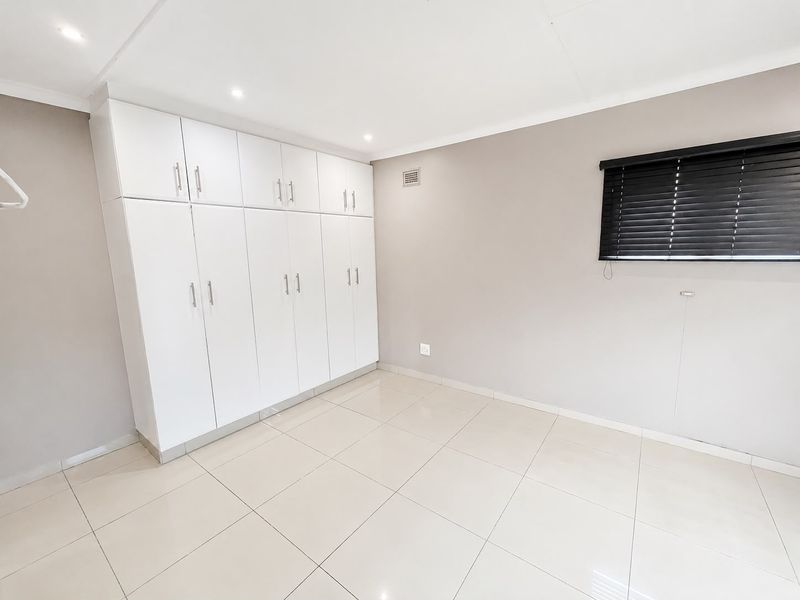 A bright, empty bedroom featuring neutral tiled flooring, modern built-in cupboards, and dark venetian blinds.