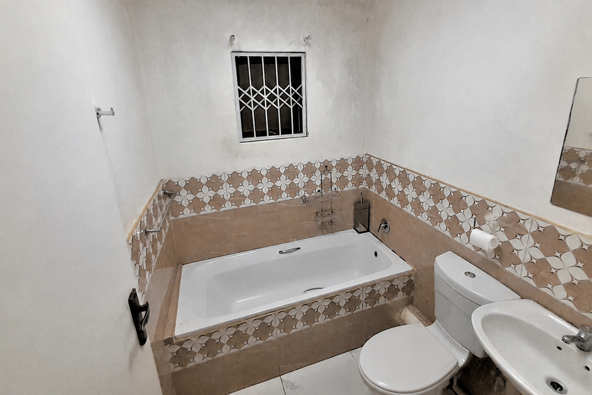 A compact bathroom featuring a built-in bathtub, pedestal washbasin, and toilet, finished with patterned ceramic wall tiles and security bars on the window.