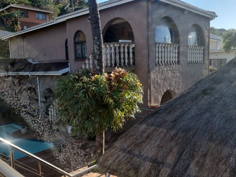 Rear view of a multi-story house featuring a swimming pool, upper-level balcony with balustrades, and a thatched roof structure in the foreground.