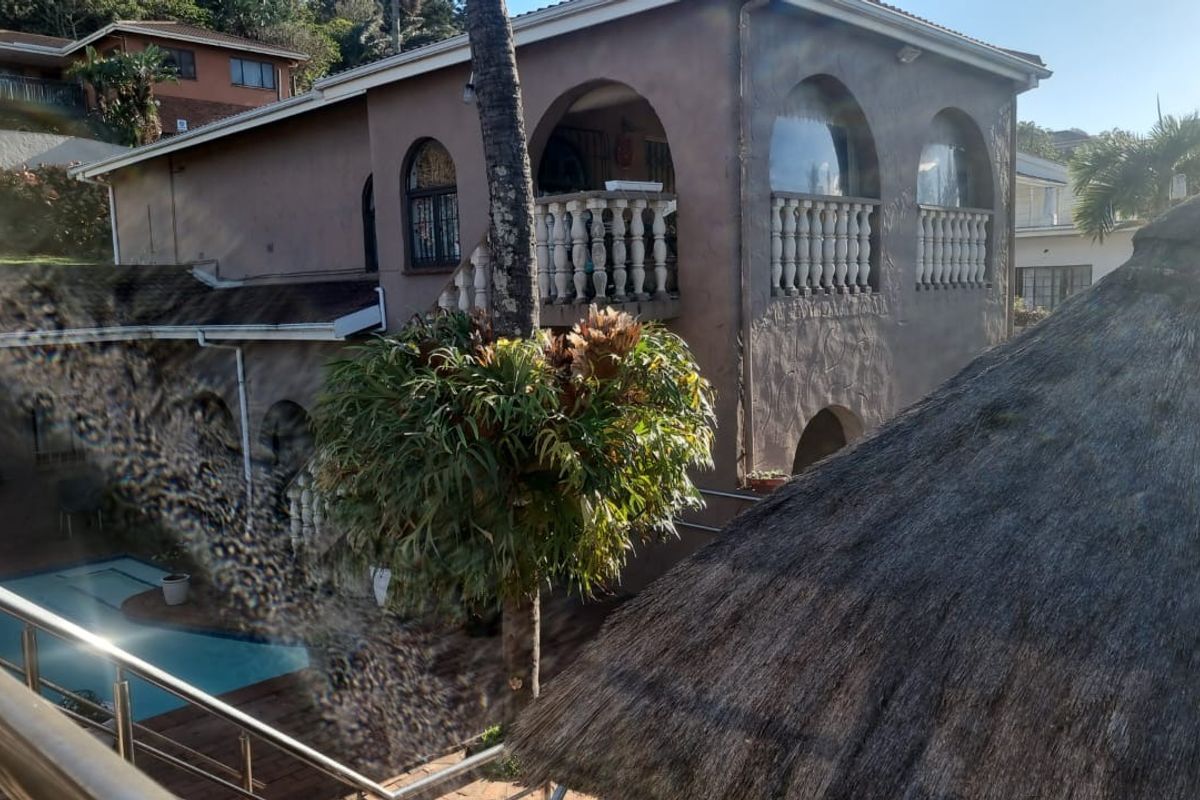Rear view of a multi-story house featuring a swimming pool, upper-level balcony with balustrades, and a thatched roof structure in the foreground.