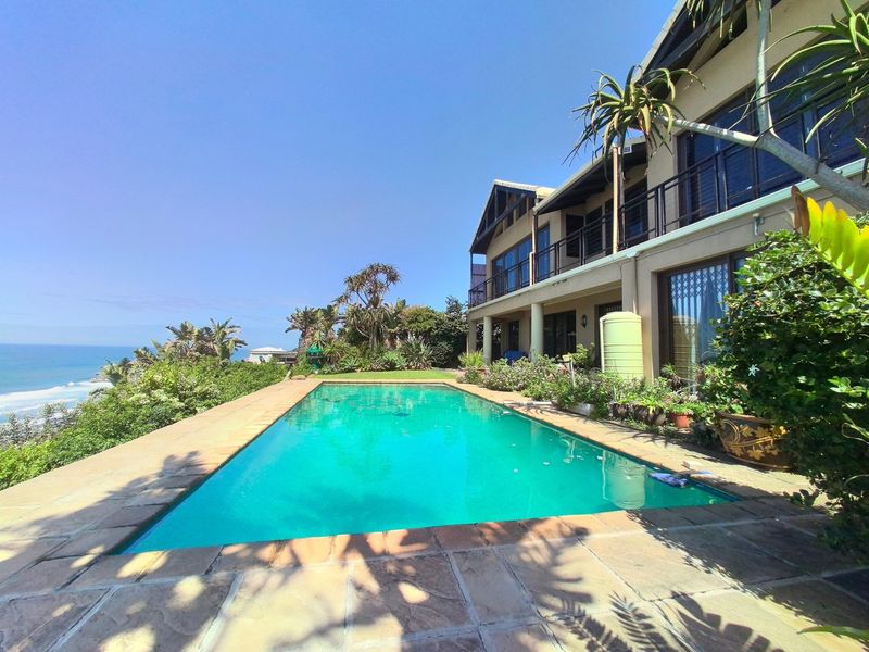 A spacious outdoor pool area featuring stone paving, overlooking the ocean with a multi-story home and balcony in the background.
