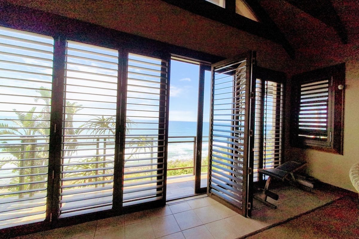 Spacious living area featuring vaulted ceilings, tiled floors, and large timber shutters opening onto a balcony with expansive ocean views.