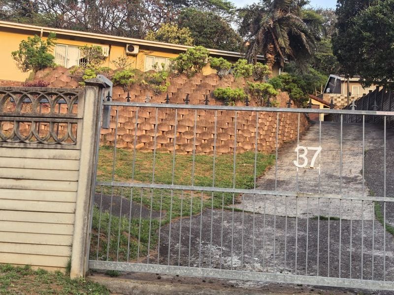 Front view of the property featuring a concrete driveway, metal security gate, and a tiered retaining wall leading up to the main house structure.