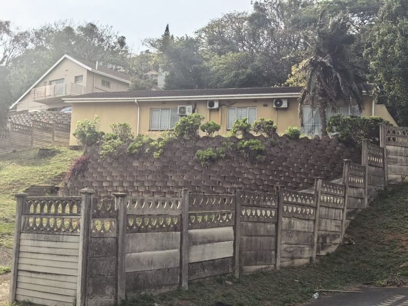 Front exterior view of a split-level house featuring a prominent retaining wall, precast concrete boundary fencing, and established garden surroundings.