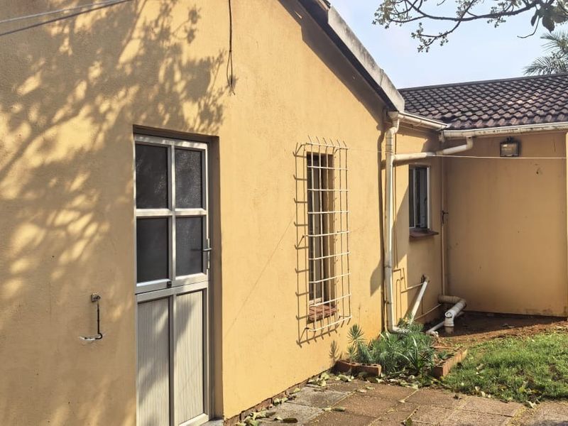 Rear exterior view of the house showing yellow plastered walls, security bars on windows, tiled roof, and a paved area with a retaining wall.