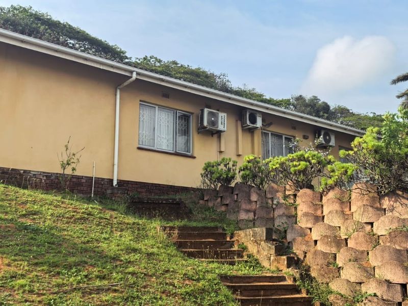 Rear exterior view of the house featuring a sloped garden, concrete retaining wall, and external air conditioning units mounted on the wall.