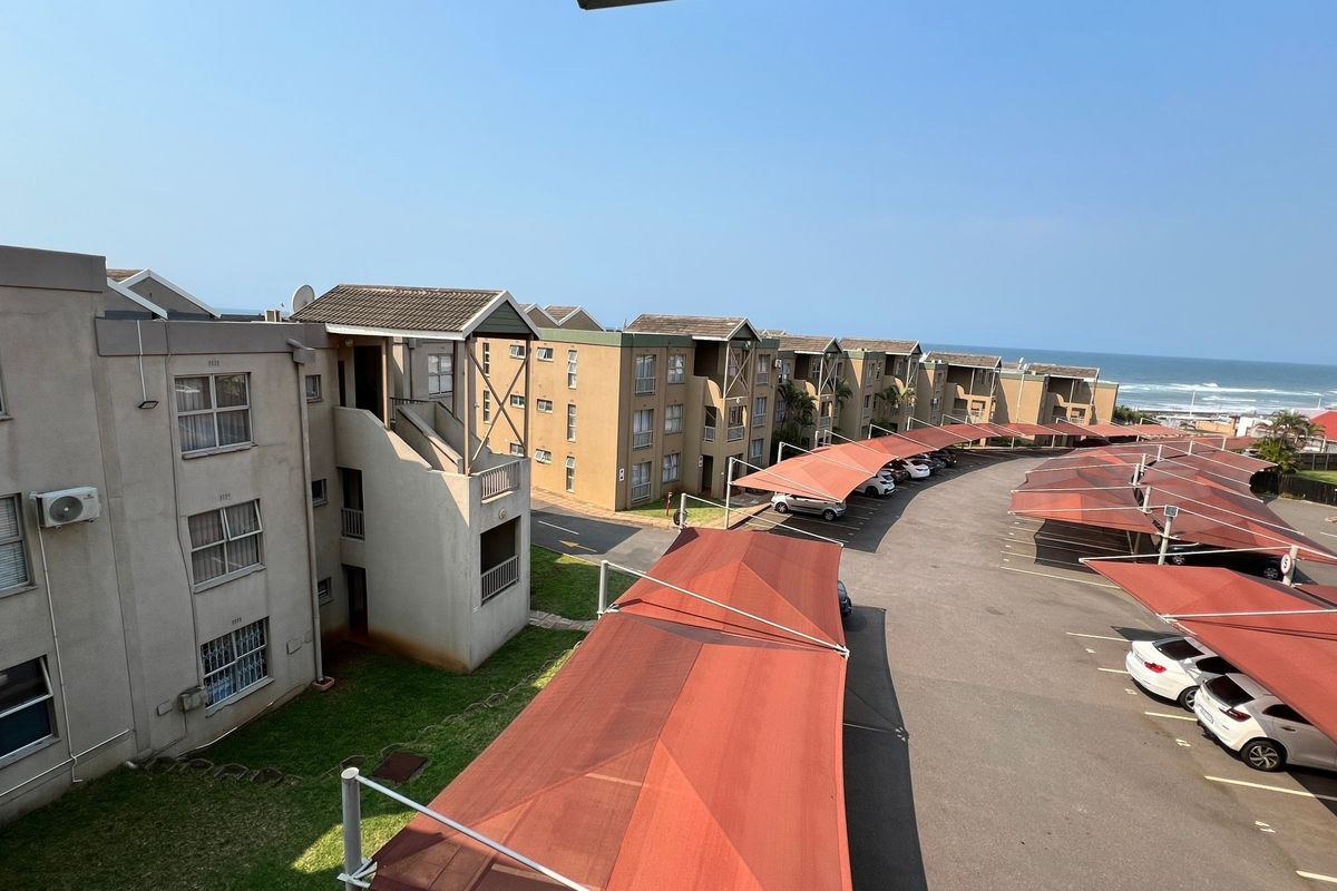 Aerial view of the apartment complex featuring covered parking bays, paved driveways, and exterior building facades with ocean views in the distance.