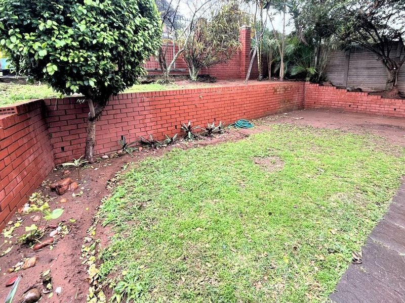 A private rear garden featuring a red brick retaining wall, established trees, and a paved walkway area.