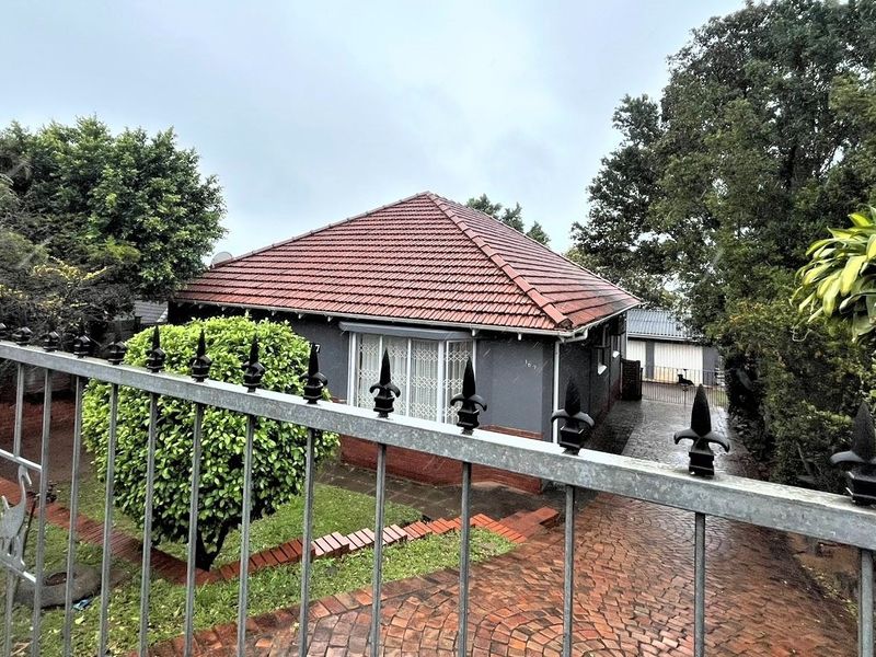 Front view of a detached house featuring a tiled roof, brick driveway, and secure metal fencing, set within a landscaped garden area.