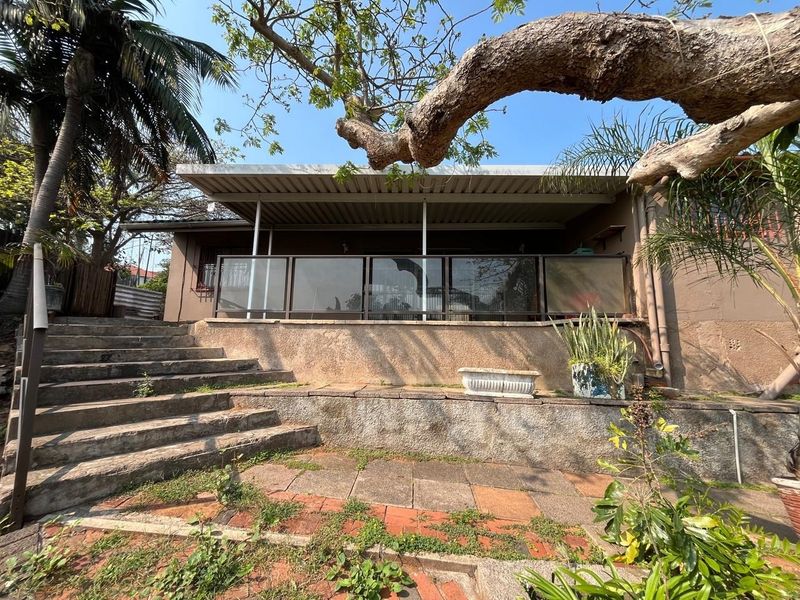 Rear exterior view featuring a covered patio with glass balustrades, concrete stairs, and a paved terrace area set within a garden.