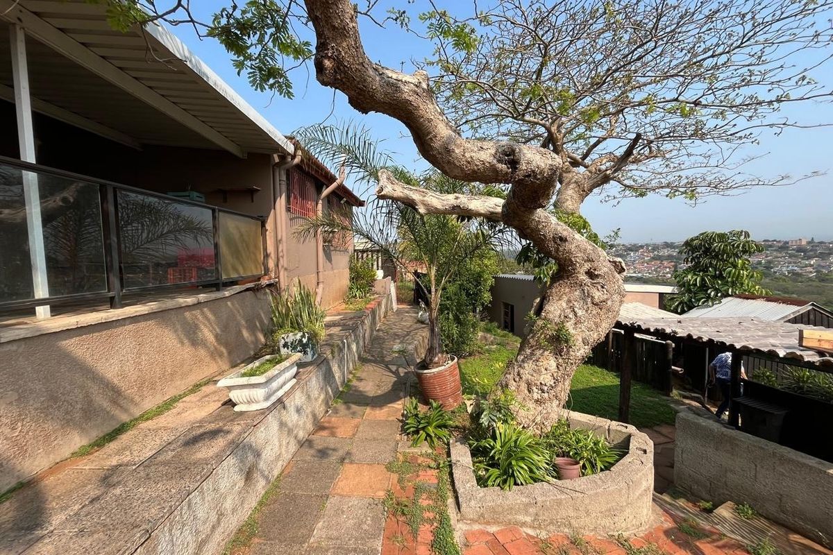Rear exterior area featuring a paved walkway, raised planter beds, and a covered patio space with a glass balustrade overlooking the surrounding neighborhood.