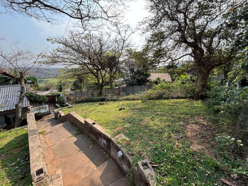 A sloped garden area featuring concrete stairs, retaining walls, and mature trees with views of the surrounding neighborhood.