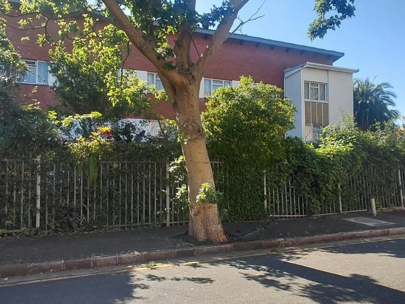 Street view of the apartment building exterior featuring a brick facade, metal perimeter fencing, and mature trees along the sidewalk.