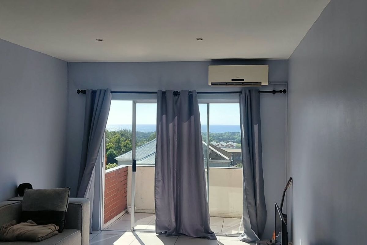 A bright living room featuring tiled floors, recessed lighting, and a split-system air conditioner, with sliding doors opening to a balcony with scenic views.