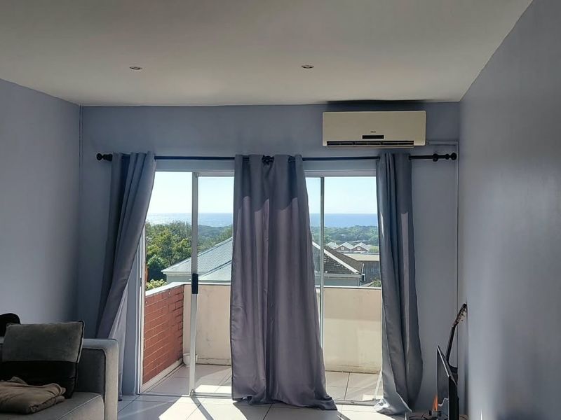 A bright living room featuring tiled floors, recessed lighting, and a split-system air conditioner, with sliding doors opening to a balcony with scenic views.