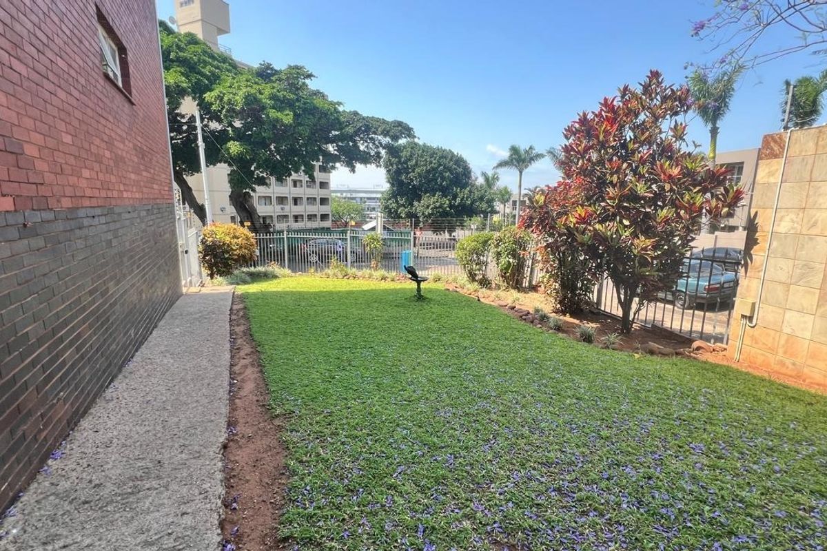 A private rear garden area featuring a well-maintained lawn, concrete pathway, and secure perimeter fencing adjacent to the apartment building.