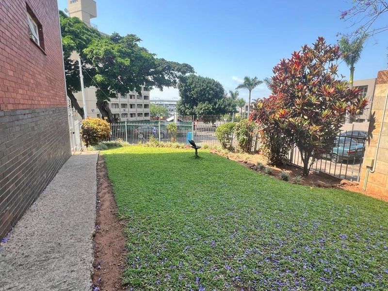 A private rear garden area featuring a well-maintained lawn, concrete pathway, and secure perimeter fencing adjacent to the apartment building.