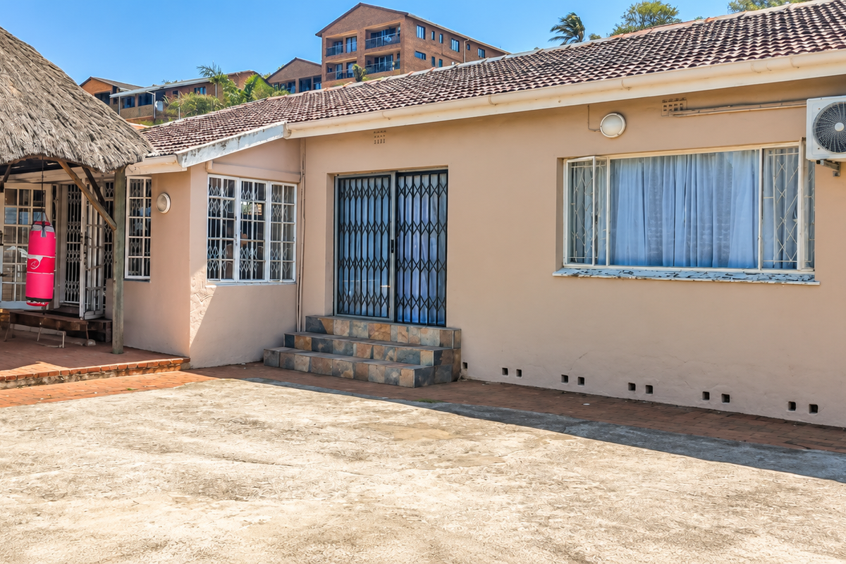 Rear exterior view featuring a thatched lapa, paved patio area, and concrete driveway, with security gates and an external air conditioning unit installed.