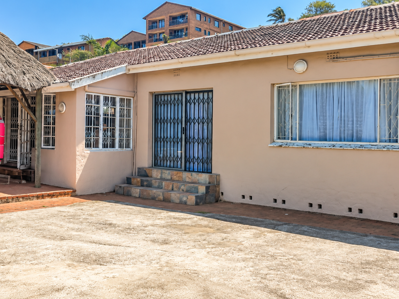 Rear exterior view featuring a thatched lapa, paved patio area, and concrete driveway, with security gates and an external air conditioning unit installed.