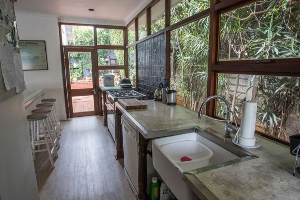 A bright kitchen featuring concrete countertops, a farmhouse sink, and extensive timber-framed windows overlooking the garden, creating a light-filled workspace.