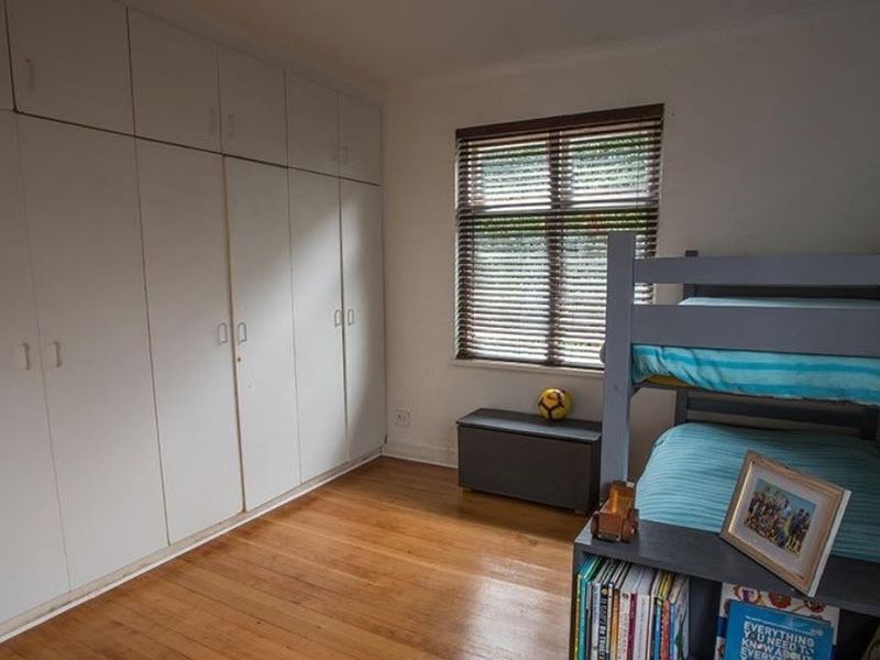 A bedroom featuring timber flooring, built-in floor-to-ceiling cupboards, and a window fitted with venetian blinds.