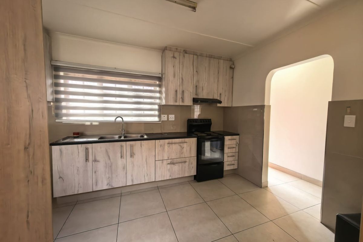 A clean, functional kitchen featuring light wood-look laminate cabinetry, tiled flooring, a stainless steel sink, and a freestanding stove with an extractor fan.