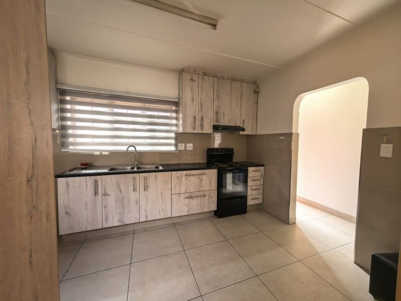 A clean, functional kitchen featuring light wood-look laminate cabinetry, tiled flooring, a stainless steel sink, and a freestanding stove with an extractor fan.