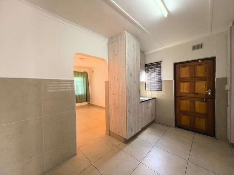 A clean, functional kitchen area featuring tiled flooring, neutral wall tiles, and modern wood-look cabinetry, leading into an adjacent living space.