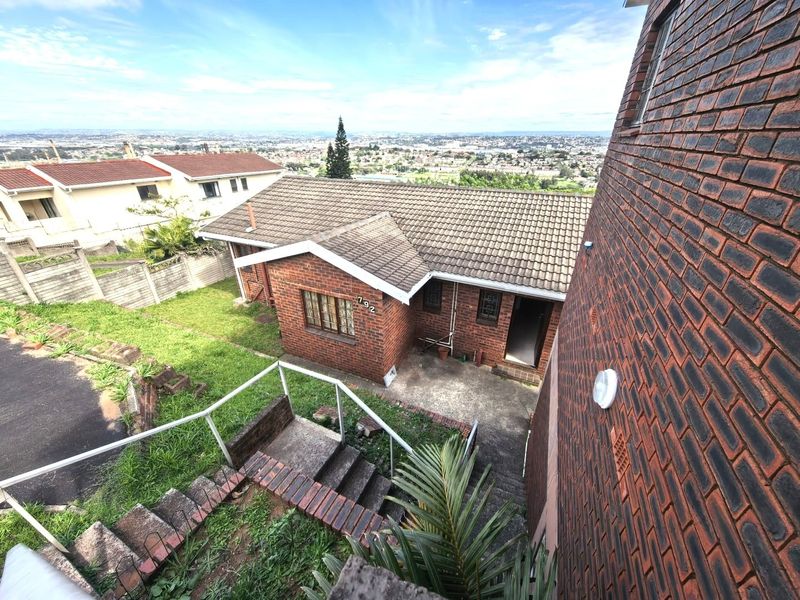 An elevated view of the rear exterior of a brick house, featuring a tiled roof, concrete staircase, and a paved entrance area.