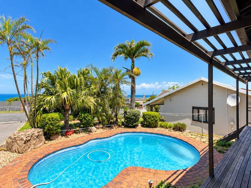 Outdoor pool area featuring a kidney-shaped swimming pool with brick coping, surrounded by a landscaped garden and a covered timber deck pergola.