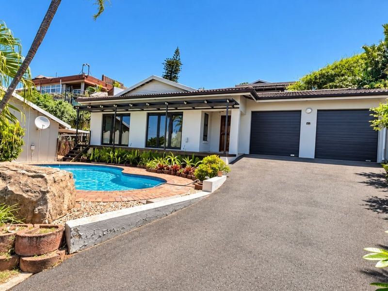 Front elevation featuring a double garage, asphalt driveway, and a swimming pool with brick coping, set within a landscaped garden area.