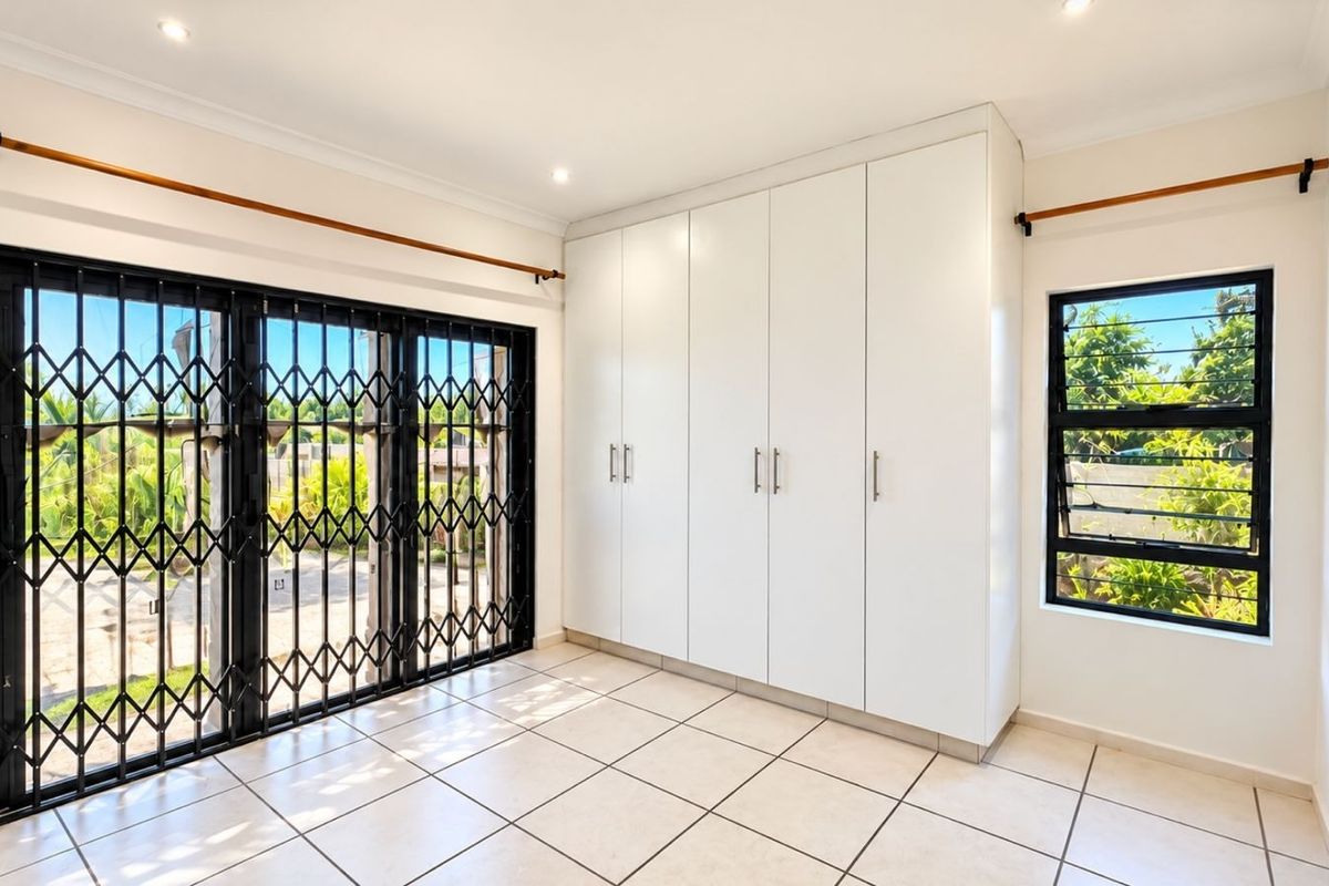 A bright, empty bedroom featuring white built-in cupboards, tiled flooring, and a large sliding door with a security gate leading to the exterior.